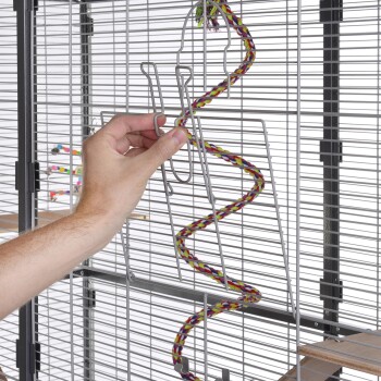 Eine Hand passt ein bunt geflochtenes Seil in einem Metallhaustierkäfig an, das die Drahtstruktur und die Holzplattform des Käfigs zeigt.