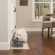 Un petit chien blanc boit dans une fontaine d'eau pour animaux grise placée sur un tapis beige dans une pièce moderne et lumineuse avec un sol en bois.