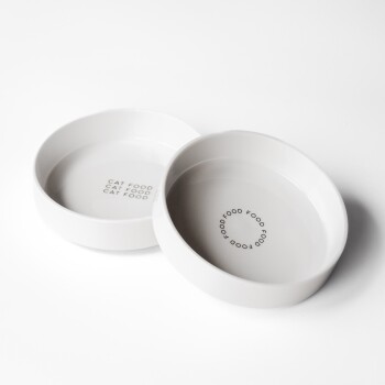 Zwei weiße Keramik-Katzenfutterschalen mit dem Text "KATZENFUTTER", der in einem kreisförmigen Muster am Boden jeder Schale aufgedruckt ist.