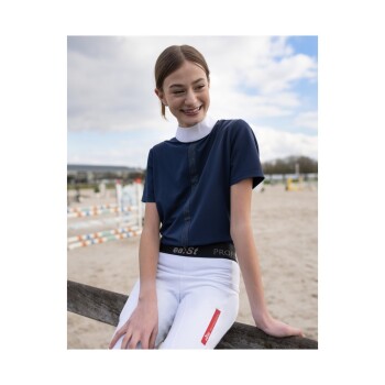 Eine junge Reiterin in einem marineblauen Reitshirt und weißen Reithosen sitzt auf einem Holzzaun in einer Pferdespringarena.