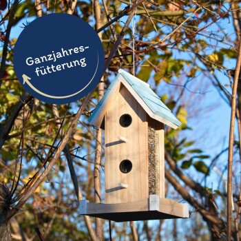 Ein Holz-Vogelfutterspender in Form eines Hauses mit einem blauen Dach, das von einem Baum hängt, mit dem Text "Ganzjahresfütterung" in einem kreisförmigen Design.