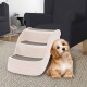 Ein kleiner Hund sitzt neben einer dreistufigen Hundetreppe in einem gemütlichen Wohnzimmer, das einen dunklen Sessel und einen hellen Teppich zeigt.