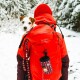 Eine Person in einer leuchtend roten Jacke und Mütze trägt einen kleinen Hund in einem Rucksack, umgeben von schneebedeckten Bäumen.
