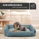 Ein kleiner Hund ruht auf einem weichen blauen Bett in einem hellen Wohnzimmer, mit dem Text: "ZUM EINKUSCHELN" und "schafft ein gemütliches Ambiente."