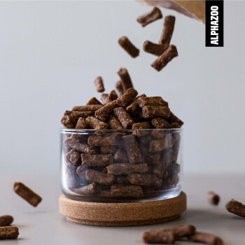 Ein Glasbehälter gefüllt mit braunen Haustierfutterpellets, mit einigen Pellets, die herausfallen, und dem Markennamen "ALPHAZOO" sichtbar in der Ecke.