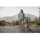 Eine Frau in einem grauen Tanktop und grünen Shorts führt einen braunen Hund auf einem Kiesweg, umgeben von Bäumen und Bergen im Hintergrund.