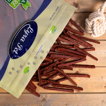 Eine Tüte mit Haustierleckereien mit der Aufschrift "Paw Treats", die lange, dünne, braune Stäbchen auf einer Holzoberfläche auskippt, mit einem Seilspielzeug in der Nähe.