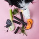 Ein schwarz-weißer Hund nähert sich einem rosa Behälter mit der Aufschrift "Darmwohl Sticks," mit dunklen, zähen Leckerlis, die teilweise sichtbar sind.