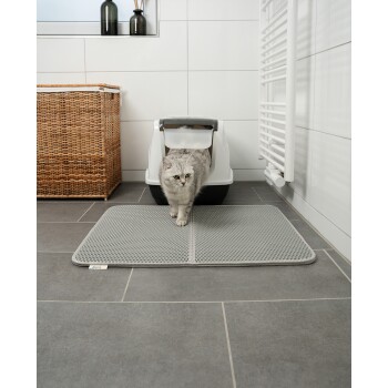 Eine graue Katze kommt aus einer weißen Katzentoilette auf eine strukturierte graue Matte in einem modernen Badezimmer mit einem Korb aus Weide und weißen Fliesen.