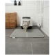 Eine graue Katze kommt aus einer weißen Katzentoilette auf eine strukturierte graue Matte in einem modernen Badezimmer mit einem Korb aus Weide und weißen Fliesen.