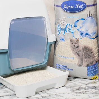 Eine Katzenstreu-Box mit einer transparenten Tür neben einem Sack Lyra Pet Graue Katzenstreu, mit einem Bild einer grauen Katze und floralem Design.