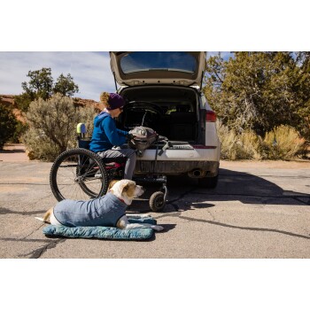 Eine Person im Rollstuhl sitzt neben einem Auto mit offenem Kofferraum, während ein Hund in einem blauen Pullover auf einer grünen Matte in der Nähe liegt.