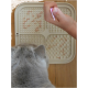 Eine graue Katze schaut auf eine beige Fressmatte mit verstreutem Futter, während eine Hand ein Stück Futter darüber hält.