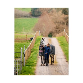 Eine Frau in einer blauen Jacke steht neben einem grauen Pferd, das eine dunkle Decke trägt, auf einem Kiesweg, umgeben von grünen Feldern und Holzzaun.