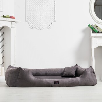 Zwei Hunde sitzen auf einem plüschigen braunen Hundebett, einer hält ein Stofftier. Der Hintergrund zeigt ein modernes Wohnzimmer mit sanfter Beleuchtung.