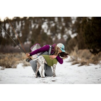 Eine Frau in einer lila Jacke und einer hellblauen Mütze kniet im Schnee und umarmt einen braun-weißen Hund, der eine grüne Weste trägt, mit Angelausrüstung in der Nähe.