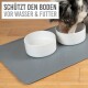 Ein Hund, der aus zwei weißen Schalen auf einer grauen Matte trinkt, mit dem Text "SCHÜTZT DEN BODEN VOR WASSER & FUTTER" darüber.