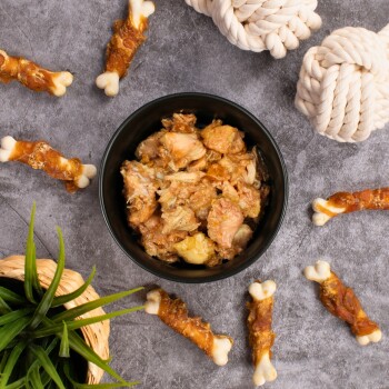 Eine schwarze Schüssel, gefüllt mit zerkleinertem Fleisch, steht auf einer grauen Oberfläche, umgeben von Hundekau-Knochen und einer kleinen grünen Pflanze in einem geflochtenen Korb.