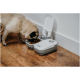 Ein Mops frisst aus einem grauen automatischen Haustierfutterspender mit offenem Deckel, der auf einem Holzboden steht.