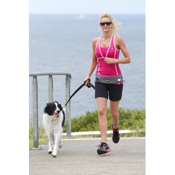 Eine Frau in einem rosa Tanktop und schwarzen Shorts joggt auf einem Weg mit einem schwarz-weißen Hund an der Leine, vor einer malerischen Ozeankulisse.