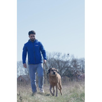 Mann in blauer Jacke mit der Aufschrift „Canine“ führt einen großen braunen Hund an der Leine durch ein grasiges Feld; im Hintergrund sind Bäume zu sehen.