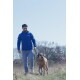 Mann in blauer Jacke mit der Aufschrift „Canine“ führt einen großen braunen Hund an der Leine durch ein grasiges Feld; im Hintergrund sind Bäume zu sehen.