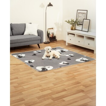 Kleiner weißer Welpe liegt auf einer grauen Haustiermatte mit schwarz-weißen Hund-Herz-Mustern in einem modernen Wohnzimmer.