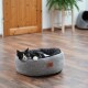 Eine schwarz-weiße Katze, die in einem gemütlichen grauen Haustierbett auf einem Holzboden entspannt, mit einer Holztür und einer Pflanze im Hintergrund.