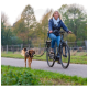 Eine Frau fährt mit einem Fahrrad einen Weg entlang, mit einem braunen Hund an der Leine neben ihr, umgeben von Grün und Bäumen.
