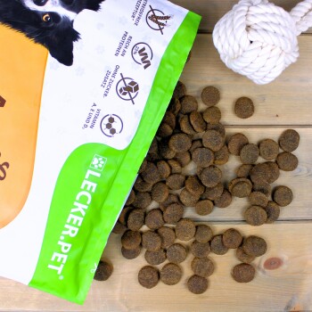 Teilweise geöffnete LECKER.PET Hundefuttertüte mit braunem Futter, das auf Holz verstreut ist, und den Hinweisen "33% Protein", "zuckerfrei" und Vitamine A, E, D.