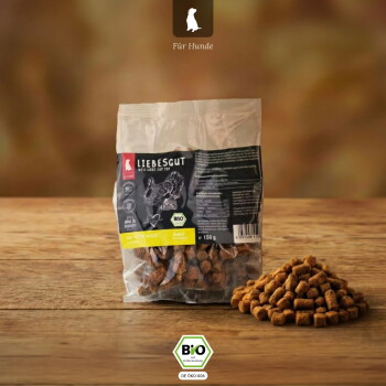 Durchsichtiger Beutel mit Liebesgut-Leckerlis (150 g) auf Holzoberfläche, Haufen Kroketten, Kopfzeile „Für Hunde“ und BIO-Sechseck (DE-ÖKO-006).