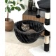 Eine graue Katze mit gelben Augen, die in einer plüschigen schwarzen Hängematte an einem Katzenbaum entspannt, mit einem Blumentopf und Holzmöbeln im Hintergrund.