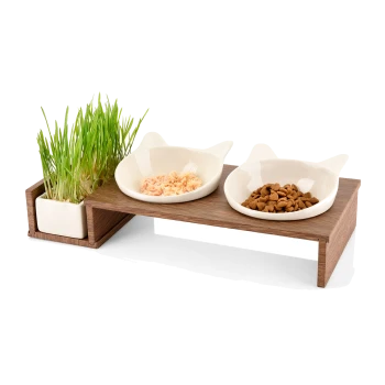 Eine hölzerne Futterstation für Haustiere mit zwei weißen katzenförmigen Schalen mit Nassfutter und Trockenfutter, plus einem kleinen Topf mit frischem Gras.