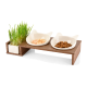 Eine hölzerne Futterstation für Haustiere mit zwei weißen katzenförmigen Schalen mit Nassfutter und Trockenfutter, plus einem kleinen Topf mit frischem Gras.