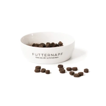 Eine weiße Futterschüssel für Haustiere, gefüllt mit dunkelbraunem Futter, mit dem sichtbaren Text "FUTTERNAPF lass es dir schmecken" und verstreutem Futter darum herum.