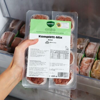 Hand hält verpackte BARFER'S Complete 'Komplett‑Mix Rind' Rohfutter-Schale (400 g) im Gefrierschrank.