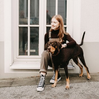 Eine Frau sitzt auf dem Boden neben einem braunen Hund, der einen gestrickten Pullover trägt, mit einem Fenster im Hintergrund.