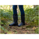 Eine Person, die stilvolle schwarze Gummistiefel mit einem gemusterten Schaft trägt, steht auf einem Waldboden, umgeben von Farnen und Laub.