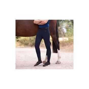 Eine Person in einem blauen Hemd und dunklen Reithosen steht neben einem braunen Pferd, wobei das Bein des Pferdes im Vordergrund sichtbar ist.