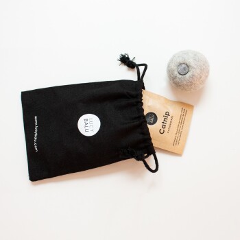 Ein schwarzer Beutel mit Kordelzug mit der Aufschrift "LUCY BALU" enthält ein braunes Paket mit der Aufschrift "Catnip" und einen grauen Filzball-Spielzeug.