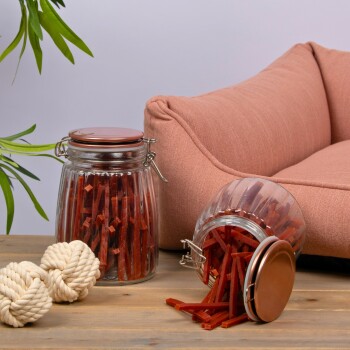 Ein Glasbehälter voller roter Haustierleckereien, der auf einen Holztisch neben einem rosa Haustierbett und einem weißen Seilspielzeug kippt.