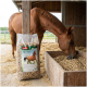 Ein braunes Pferd mit einer weißen Blässe frisst aus einer weißen Schüssel auf dem Boden, neben einem Sack PANTO Apfel-Müsli Pferdefutter.