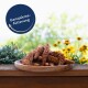 Ein Holzbrett mit strukturierten braunen Haustierfuttersticks, umgeben von grünen Pflanzen und gelben Blumen, mit einem blauen Etikett "Ganzjahresfütterung."