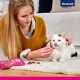 Eine Frau in einem gelben Pullover füttert eine weiße Katze mit schwarzen Markierungen Leckerlis aus einer rosa Vitakraft-Tüte auf einem beigen Teppich.