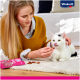 Eine Frau in einem gelben Pullover füttert eine weiße Katze mit schwarzen Markierungen Leckerlis aus einer rosa Vitakraft-Tüte auf einem beigen Teppich.