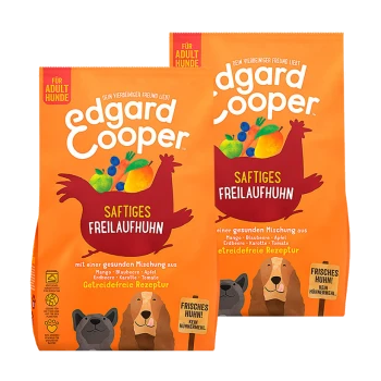 Twee zakken Edgard Cooper hondenvoer met een oranje ontwerp, cartoonkip, fruit en tekst 'Saftiges Freilaufhuhn' en 'Für Adult Hunde.'