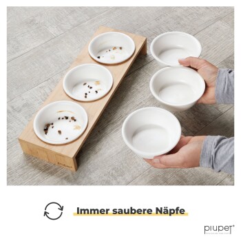 Eine Person hält drei weiße Haustiernäpfe, während sie sie auf einen Holzständer mit drei leeren Näpfen stellt, mit dem Text "Immer saubere Näpfe."