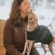 Eine Frau in einem braunen Pullover sitzt mit einem kleinen Hund, der einen dunklen Strickpullover und eine Leine trägt, beide schauen sich an.