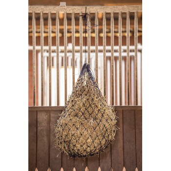 Ein Heunetz, gefüllt mit Heu, hängt an einem Metallhaken in einem Stall, mit Holzwänden und vertikalen Metallstangen im Hintergrund.