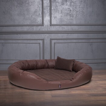 Braunes ovales Haustierbett mit erhöhten Seiten und passendem Kissen auf Holzboden vor grauer Paneelwand.